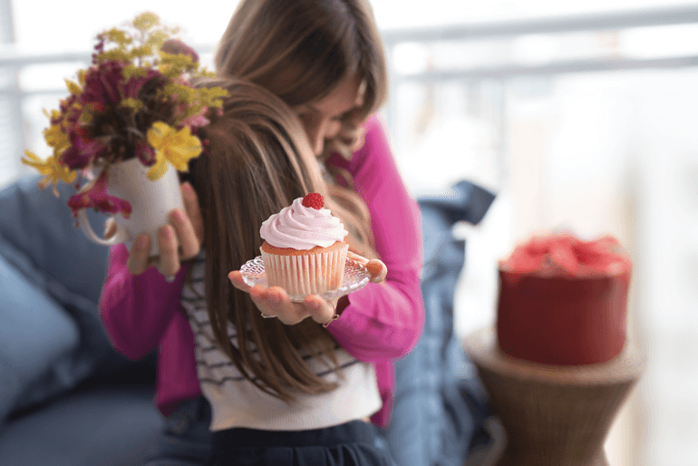 Adult woman hugging young child. White cupcake with red frosting on a plate. Chocolate cake with red frosting and white flowers on top on a plate. Wood cutting board and wooden spoon on a red table cloth. Purple and yellow flowers in vase.
