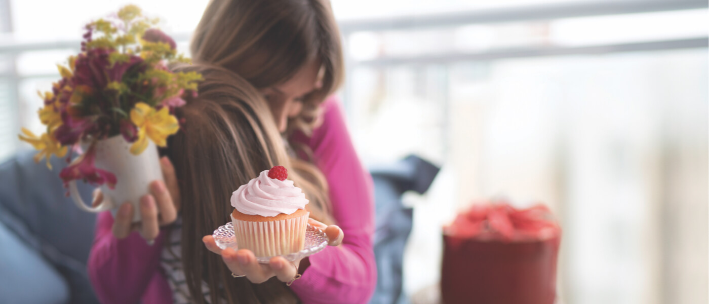 Adult woman hugging young child. White cupcake with red frosting on a plate. Chocolate cake with red frosting and white flowers on top on a plate. Wood cutting board and wooden spoon on a red table cloth. Purple and yellow flowers in vase.