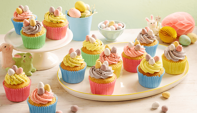 an assortment of vibrantly colored Speckled Mini Egg Cupcakes arranged on white platter, each topped with swirls of frosting & mini easter eggs